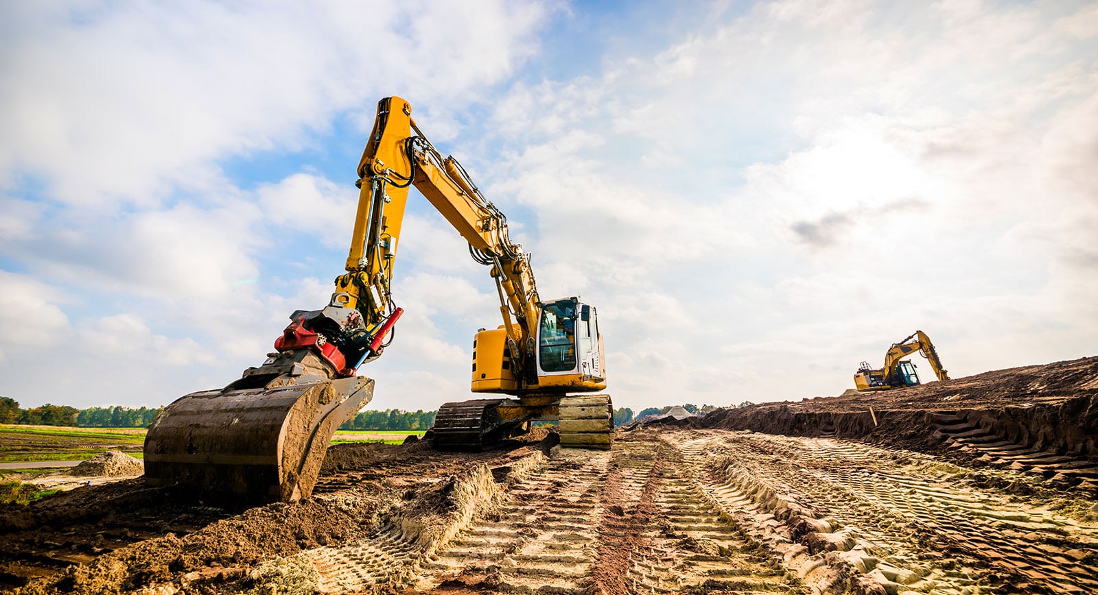 A tracked hydraulic excavator working graded earth on a construction site under a wide cloudy sky